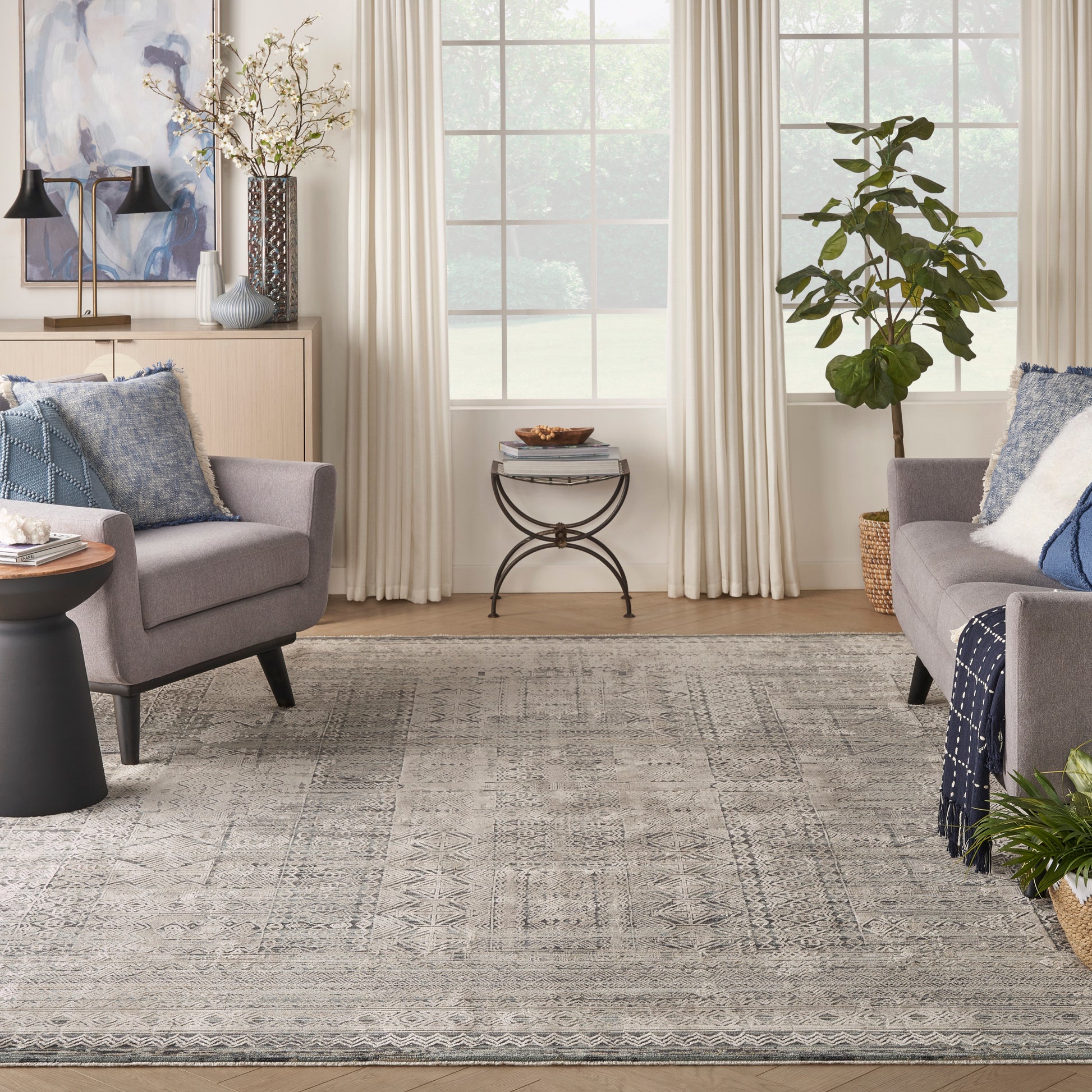 Vintage 8' x 10' rug in a cozy living room with an intricate grey pattern, two grey armchairs, blue pillows, a round metal side table and greenery.
