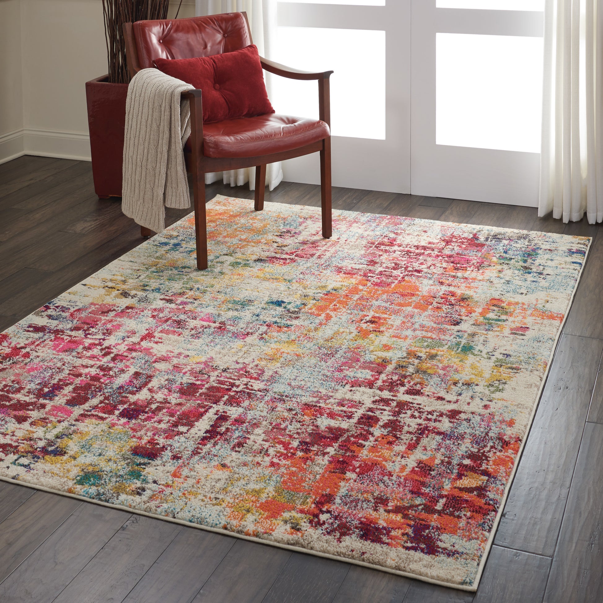 Colorful abstract area rug on dark wood floor beside red leather armchair with beige throw.
