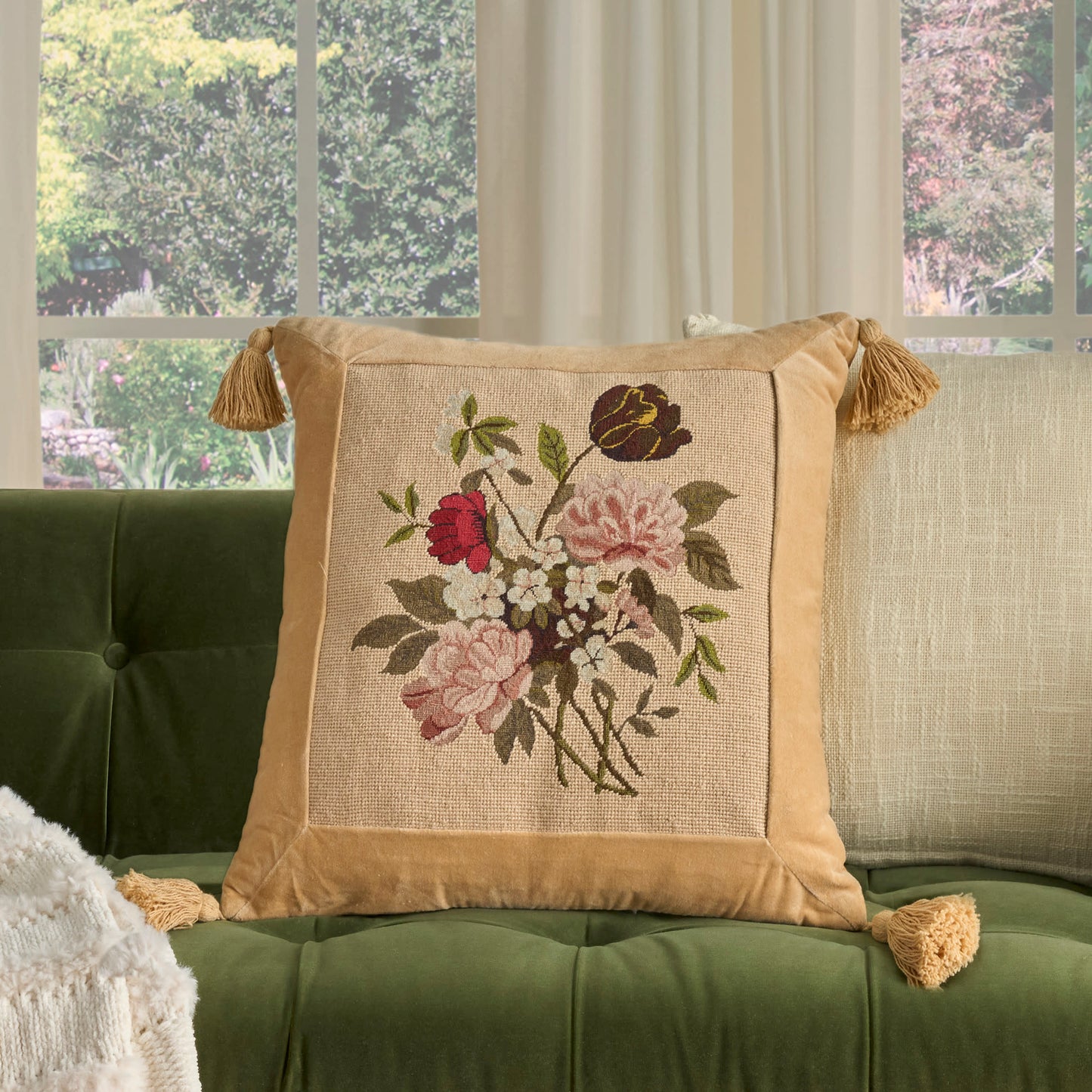 Beige square throw pillow with floral embroidery, tan satin border & tassels on corners, on a green tufted sofa.