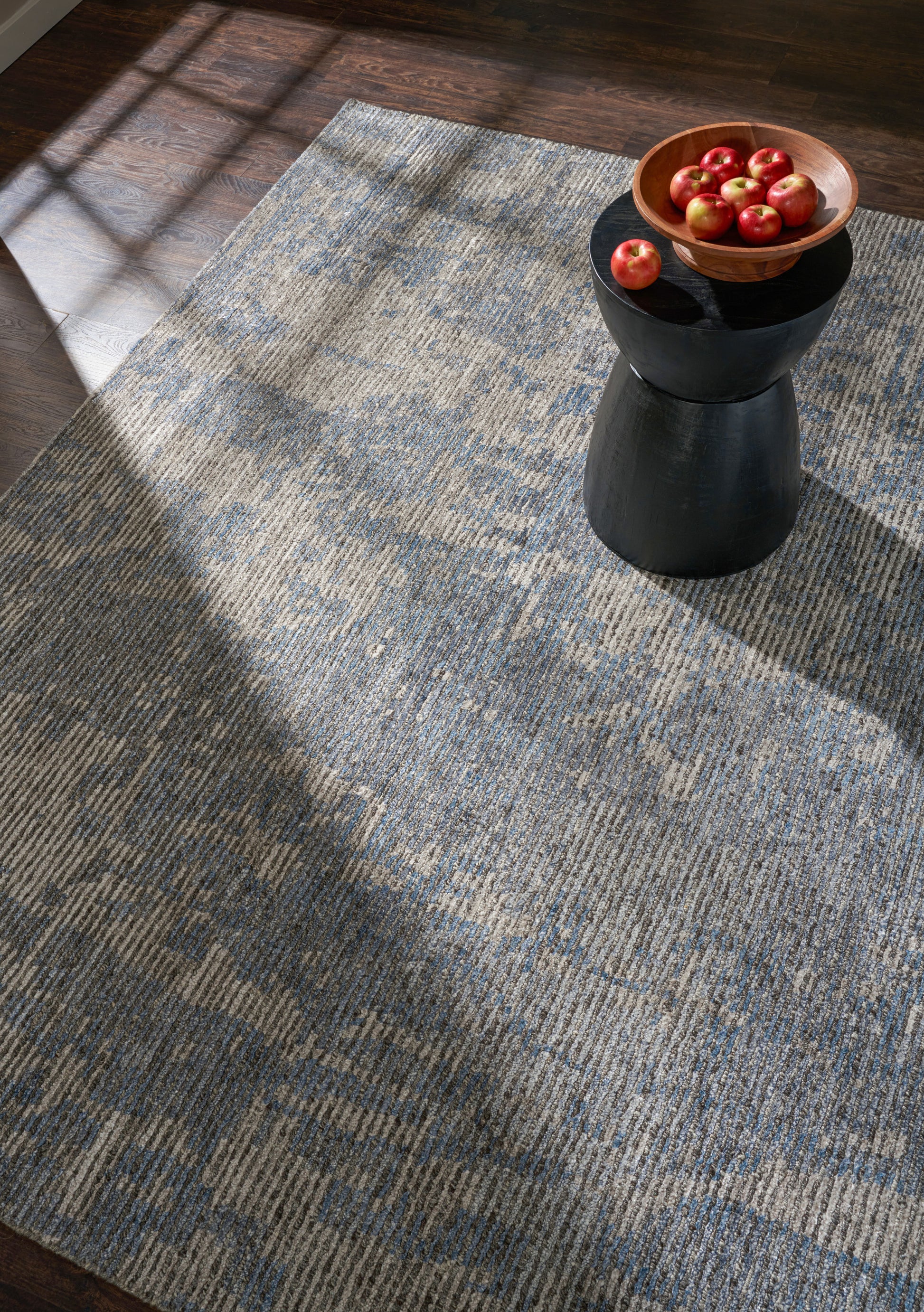Blue-gray woven rug on dark wood floor with a black pedestal holding a wooden bowl of red apples.