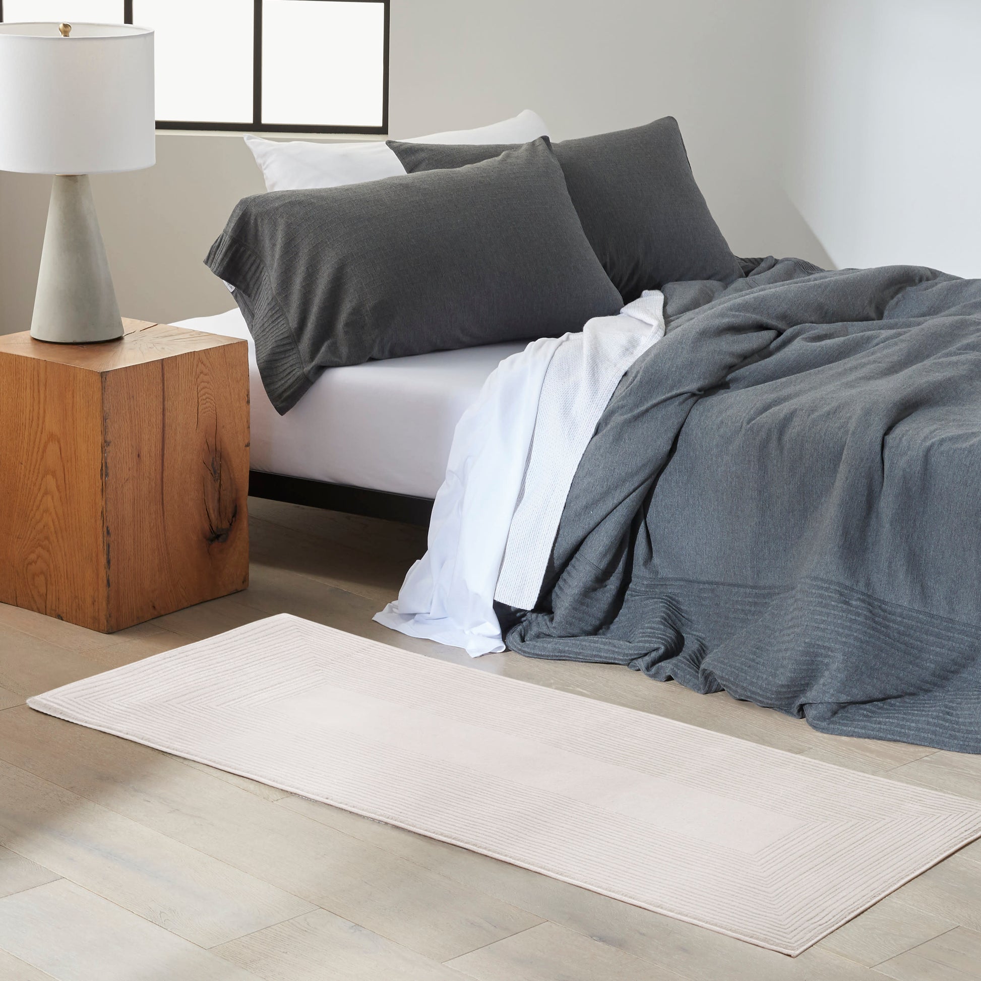 Modern bedroom: dark gray duvet and pillows on a low bed, white sheet, light beige rug, cube nightstand, ceramic lamp.