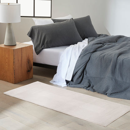 Modern bedroom: dark gray duvet and pillows on a low bed, white sheet, light beige rug, cube nightstand, ceramic lamp.
