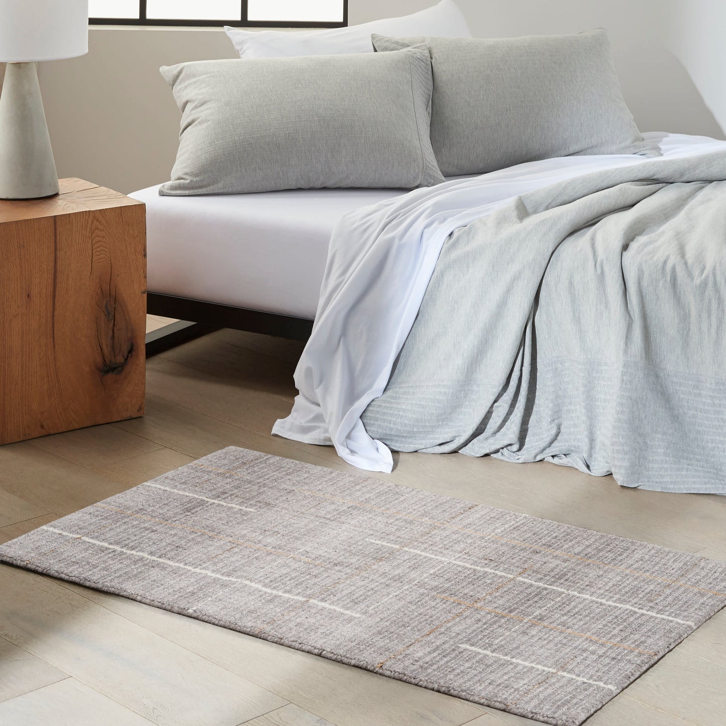 Beige-gray rectangular rug in a bedroom scene with neutral gray bedding, a light-gray throw and cushions, and a wooden nightstand.