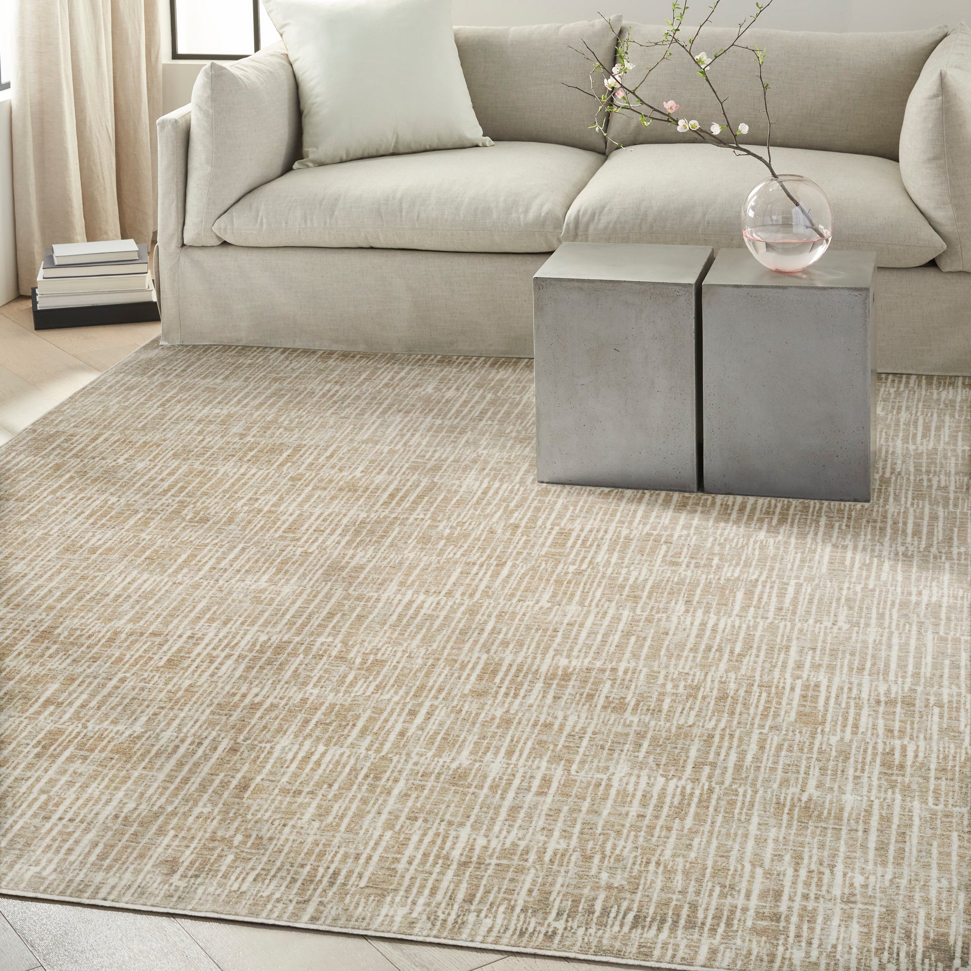 Beige fabric sofa with pale cushions, concrete block side tables, glass vase with blossoming branch, textured beige rug.