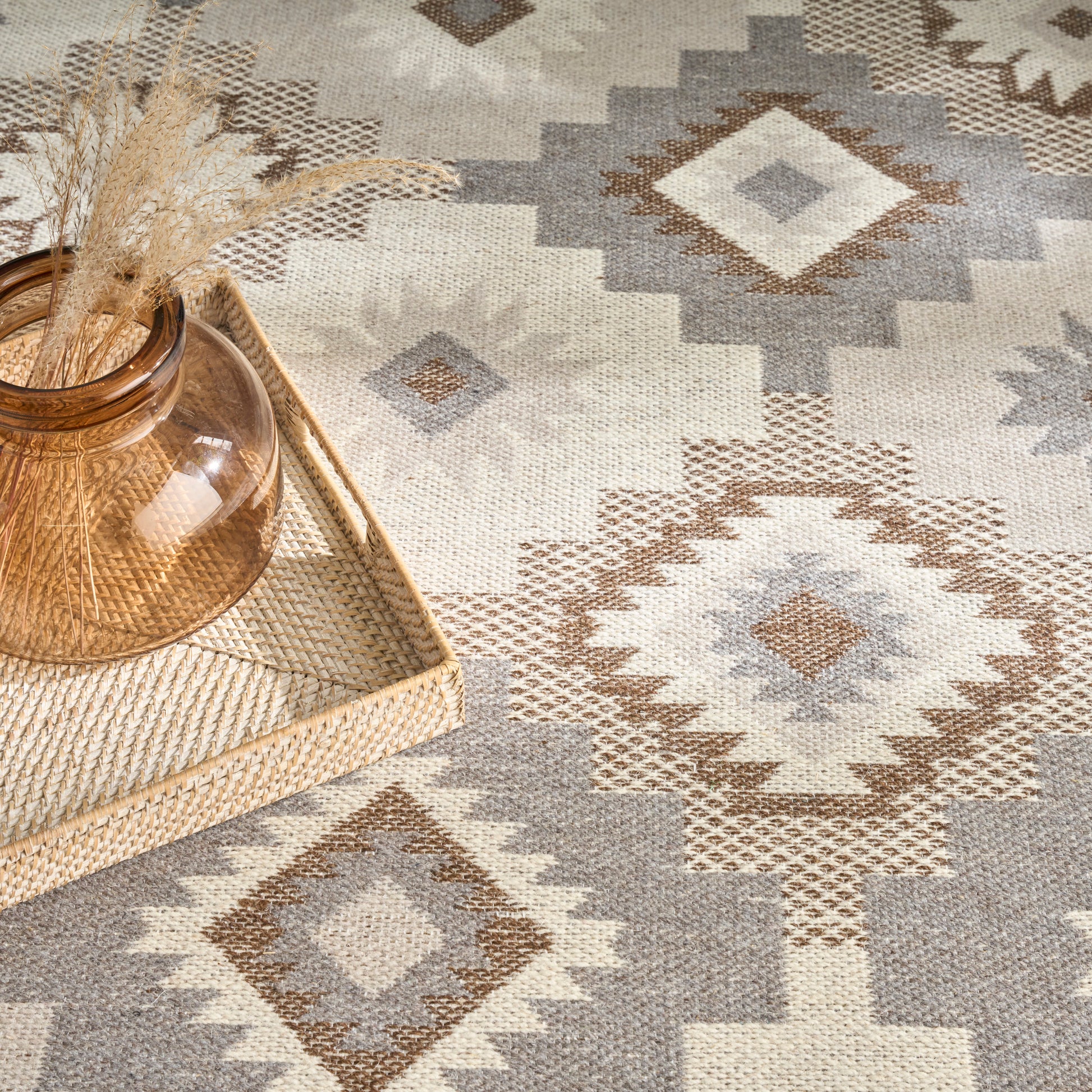 Geometric diamond-pattern rug in beige, taupe, gray with a woven tray holding an amber glass vase and dried grasses.