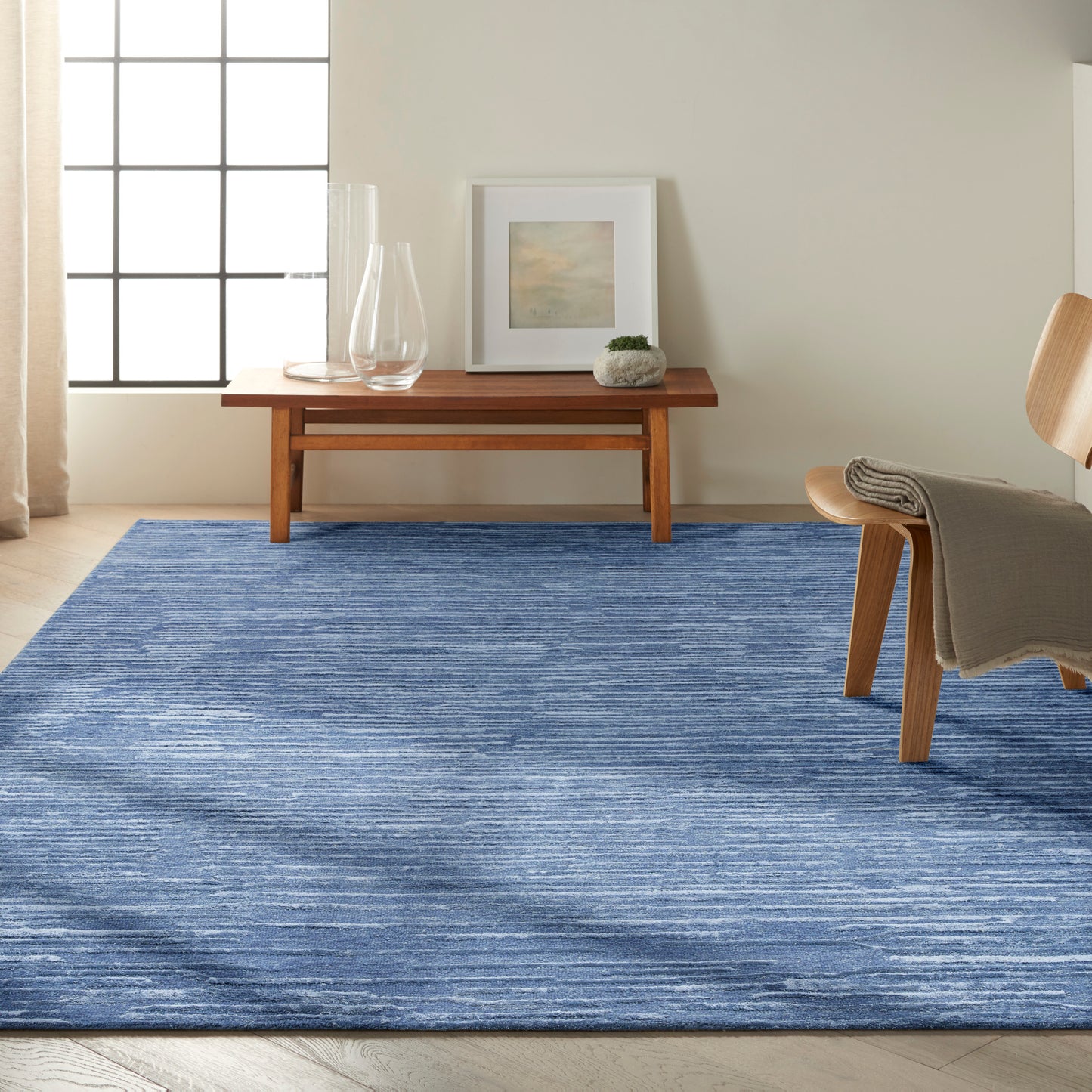 Mid-century living room with a blue textured rug, wooden coffee table, framed art, glass vases, small plant, and a wooden chair with a throw.