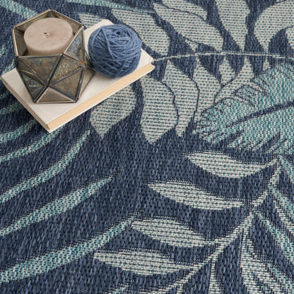 Blue and teal patterned rug with leaf motif, beige candle in geometric glass holder on a small book, and a blue yarn ball.