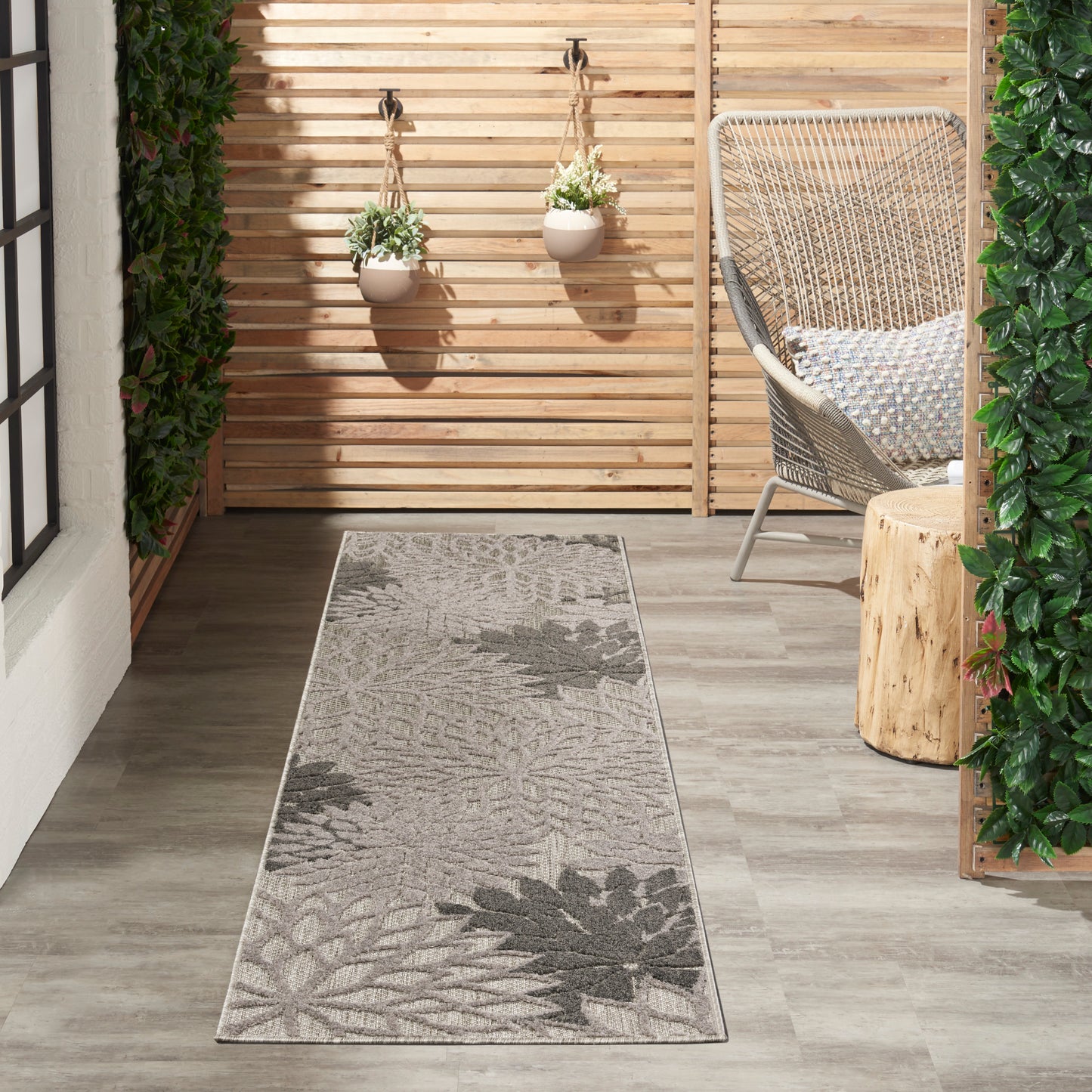 Rectangular woven rug runner in gray with botanical leaf pattern on wood floor beside hanging planters and wicker chair.