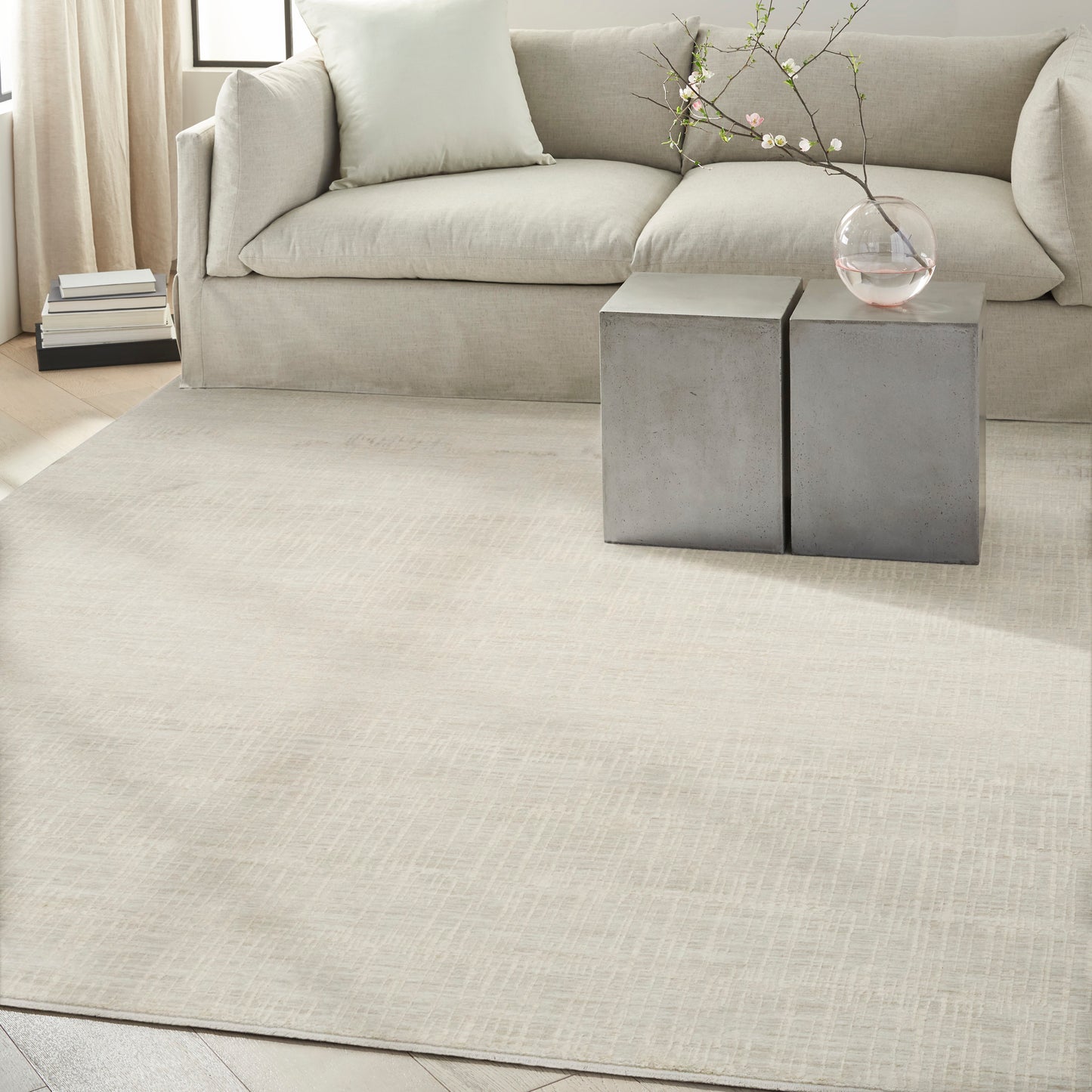 Neutral living room with beige sofa, light cream rug, two gray square side tables, glass vase with branch, and book stack.