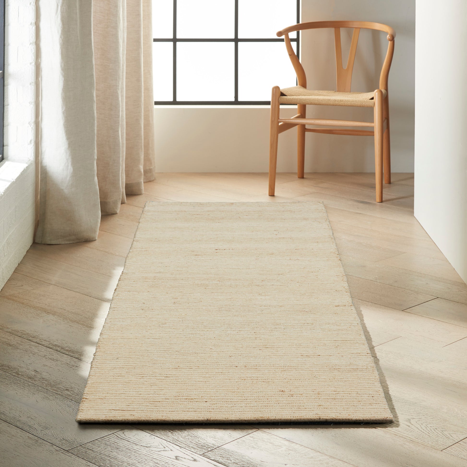 Beige woven rug runner on light wood floor beside a wooden chair with a woven seat by a large grid-window.