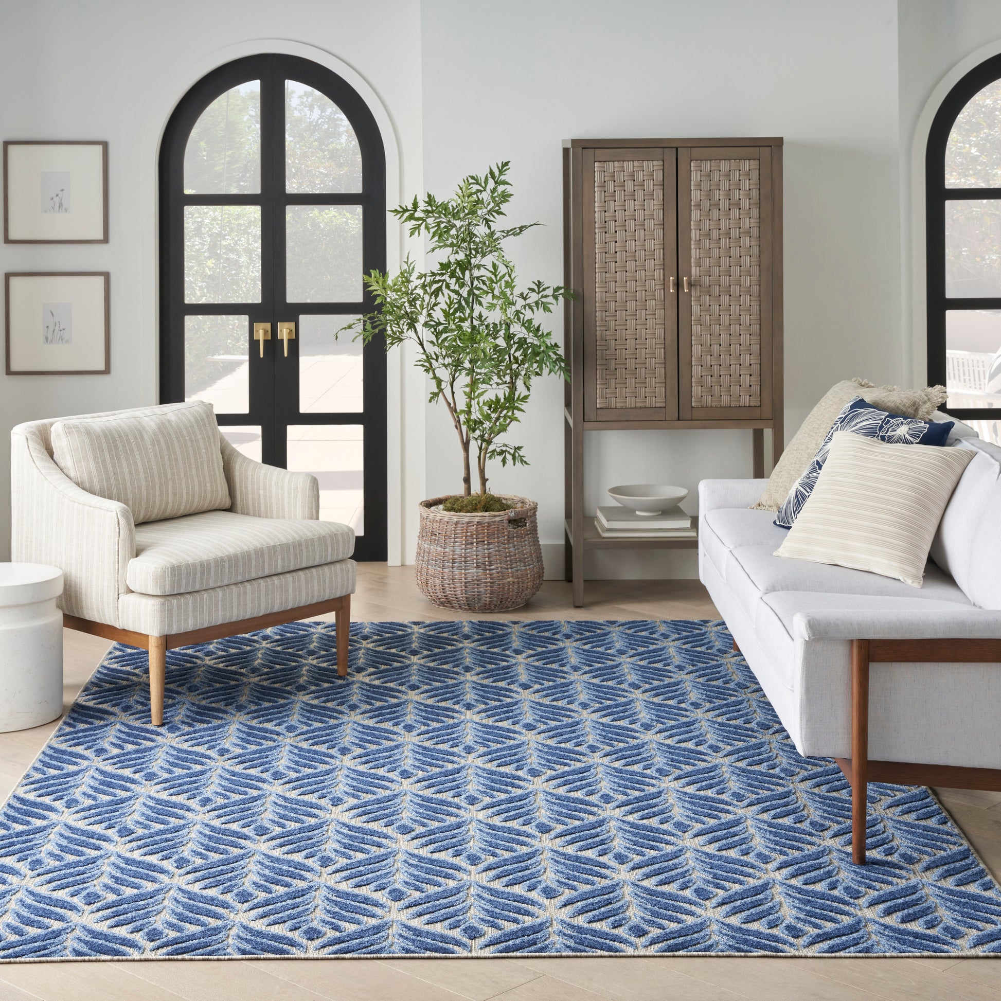 Living room with blue geometric rug, cream armchair, light sofa, rattan cabinet, potted plant, arched black doors.