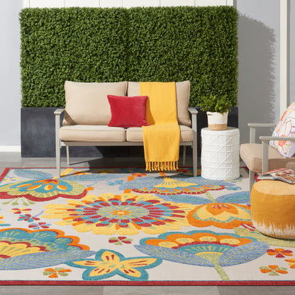 Outdoor patio with a floral rug in yellow/orange/blue; beige sofa, red pillow, mustard throw, white side table, greenery.