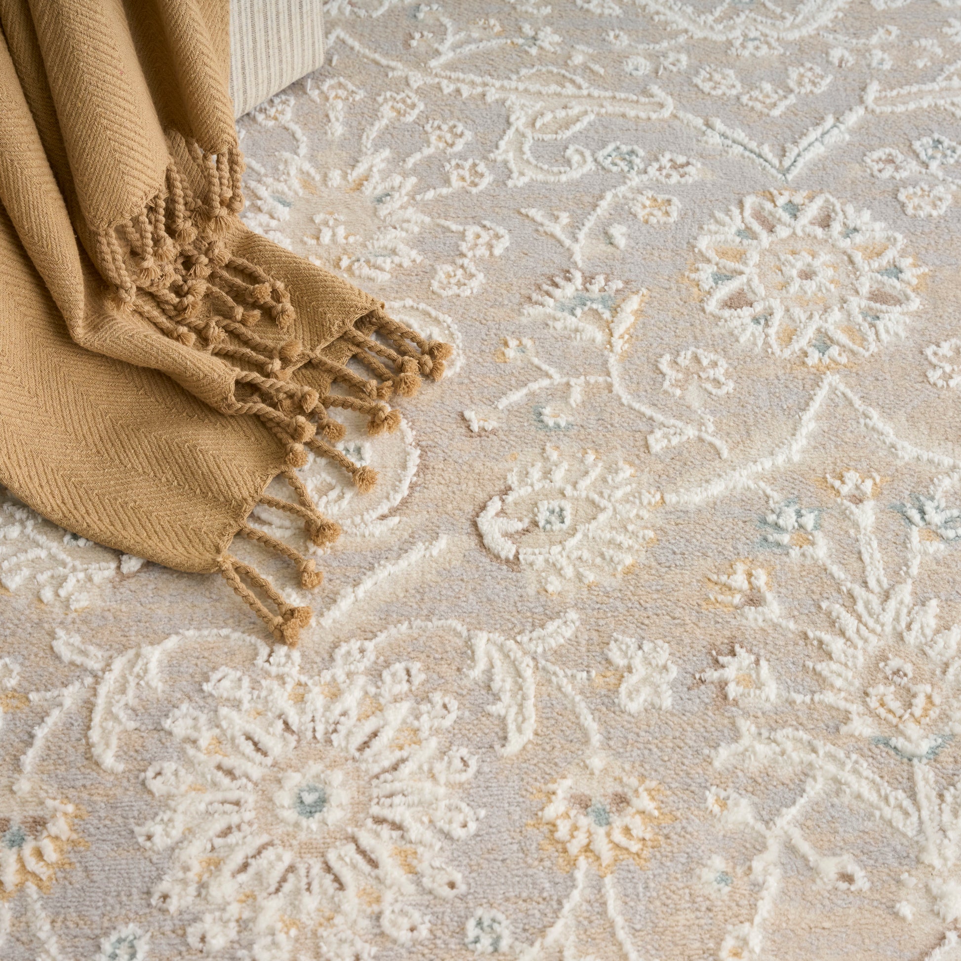 Camel knit scarf with fringed tassels draped on a beige floral rug; cozy winter accessory.