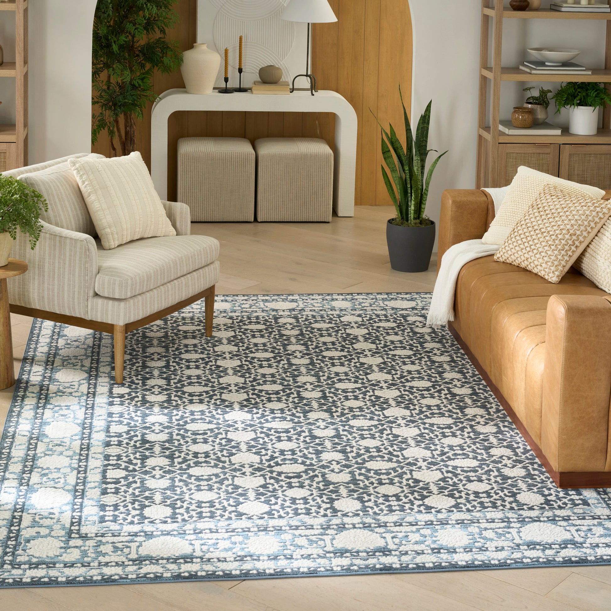 Living room with blue-and-white patterned rug, beige armchair, tan leather sofa, light wood shelves, curved white console.
