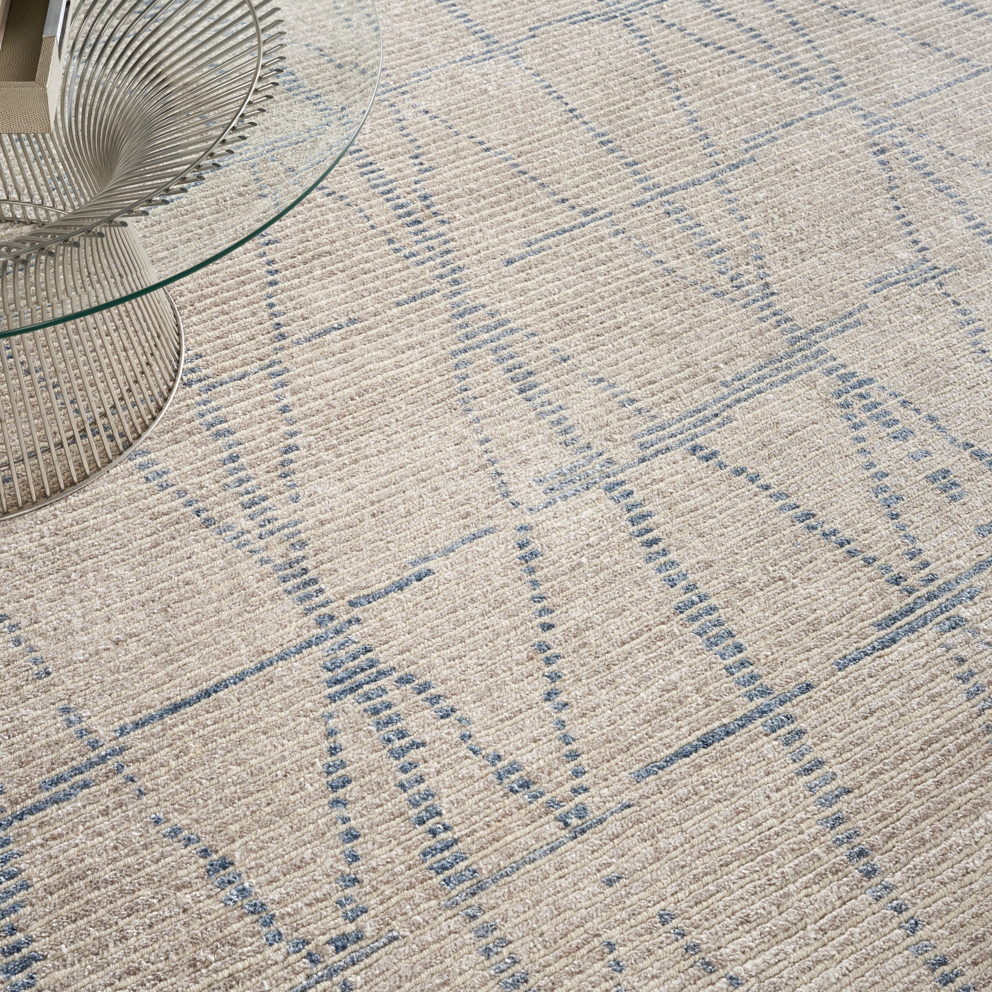 Beige textured area rug with light blue geometric grid pattern, paired with a glass round coffee table with a metal wire base.
