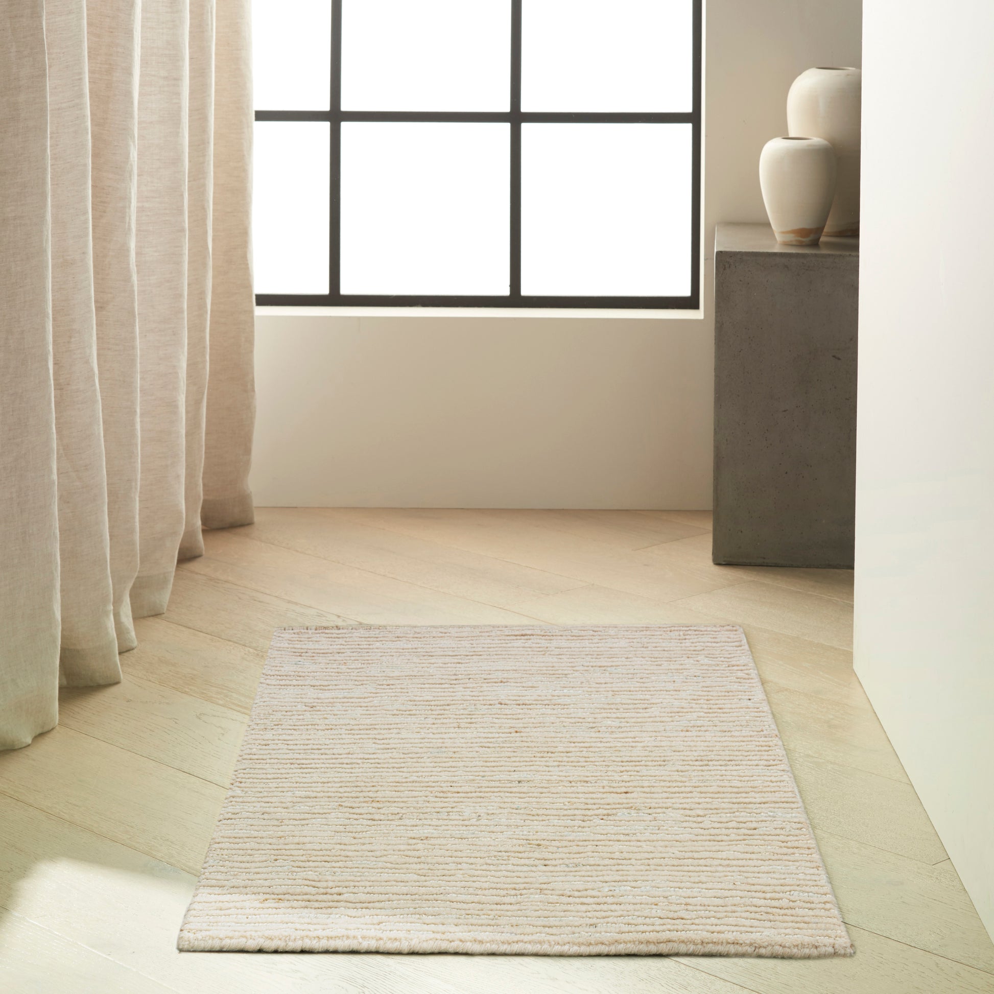 Cream ribbed rectangular rug on light wood floor beside a large window with beige curtains and a concrete pedestal with vases