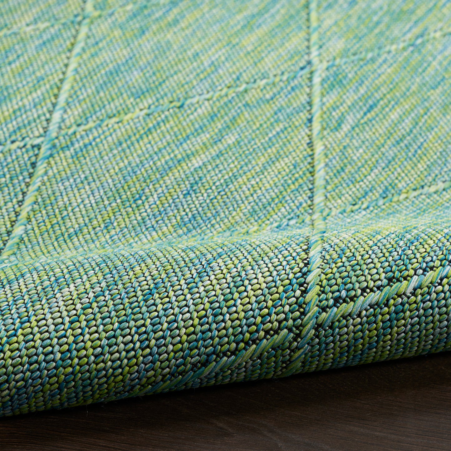 Close-up of a green-blue woven rug with quilted square grid and rolled edge on dark wood floor.