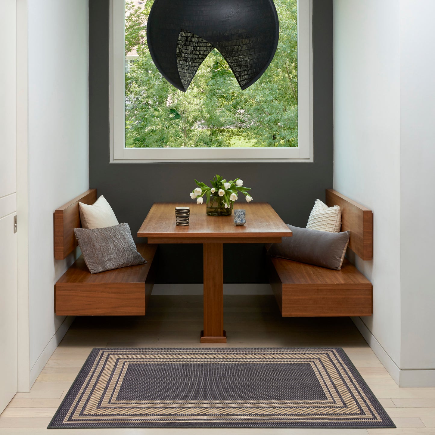 Cozy corner dining nook with wooden table and benches, gray cushions, black sculptural pendant light, greenery outside, rug.