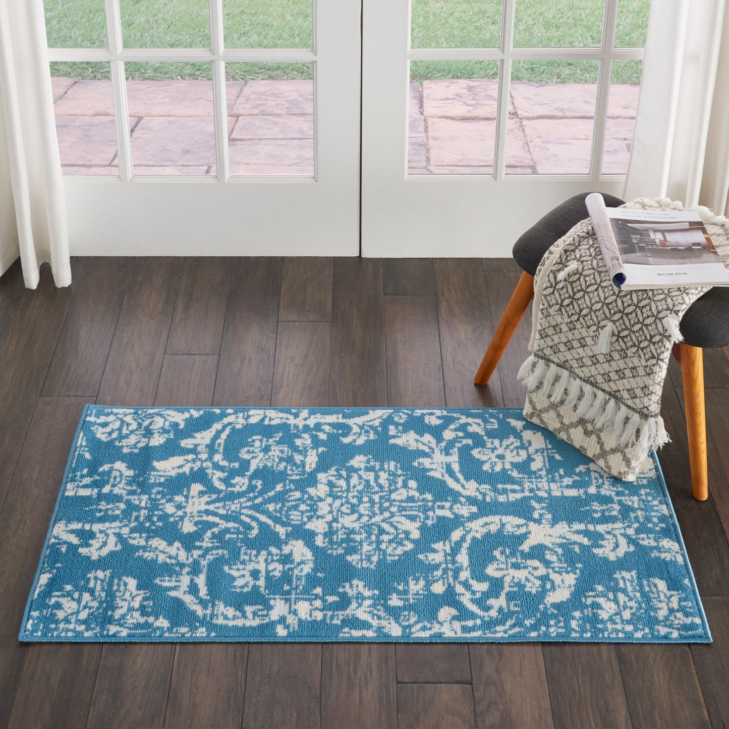 Rectangular teal rug with white damask pattern on dark wood floor, near French doors; chair with knit blanket and magazines.