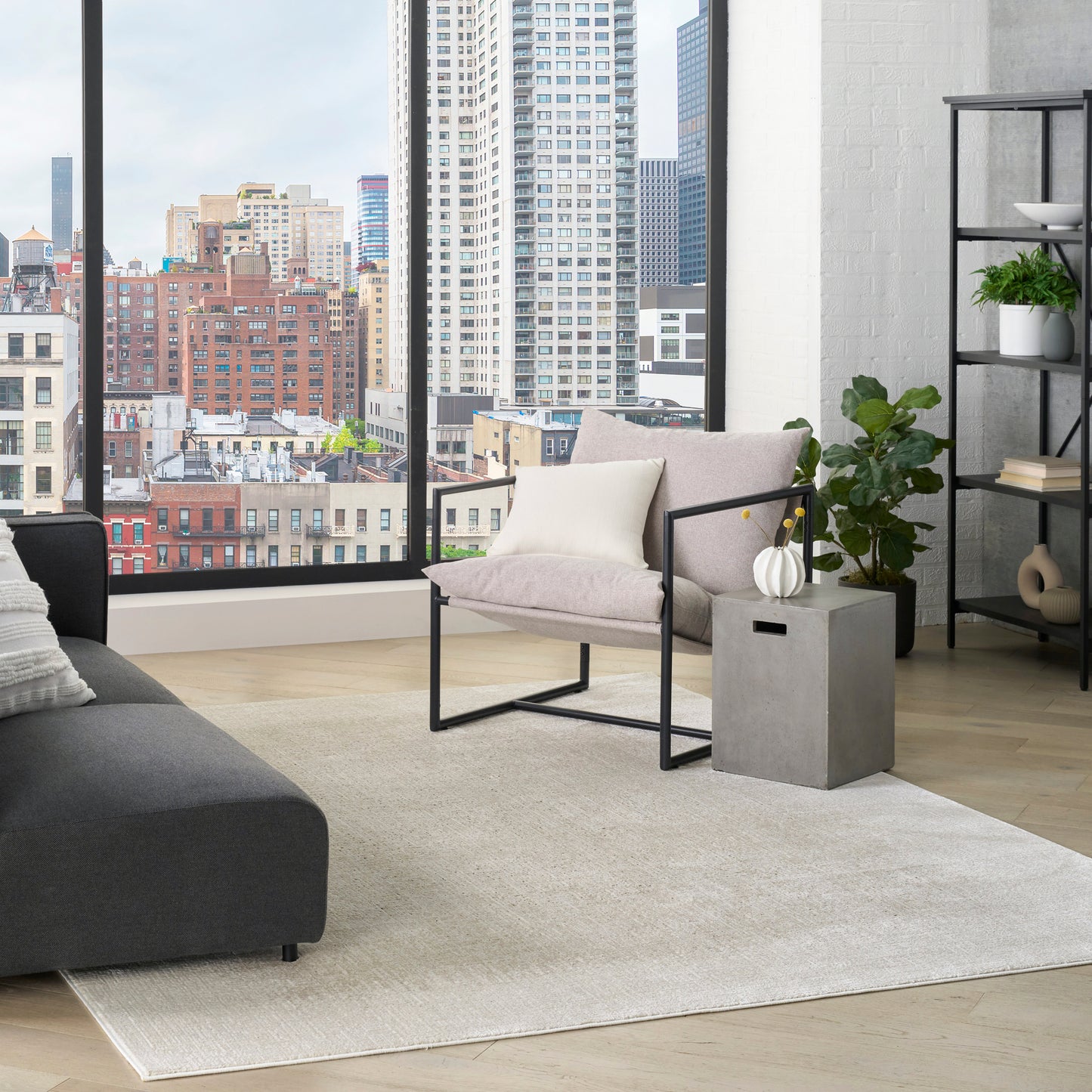 Modern living room with floor-to-ceiling city-view windows, gray sofa, beige metal-frame armchair, light rug, and plants.