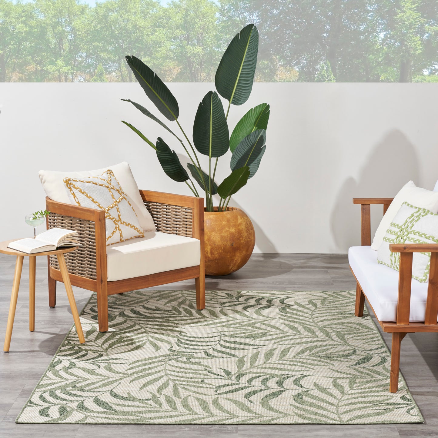 Cozy indoor patio with wood rattan chairs, white cushions, a leaf-patterned rug, and a large potted tropical plant.