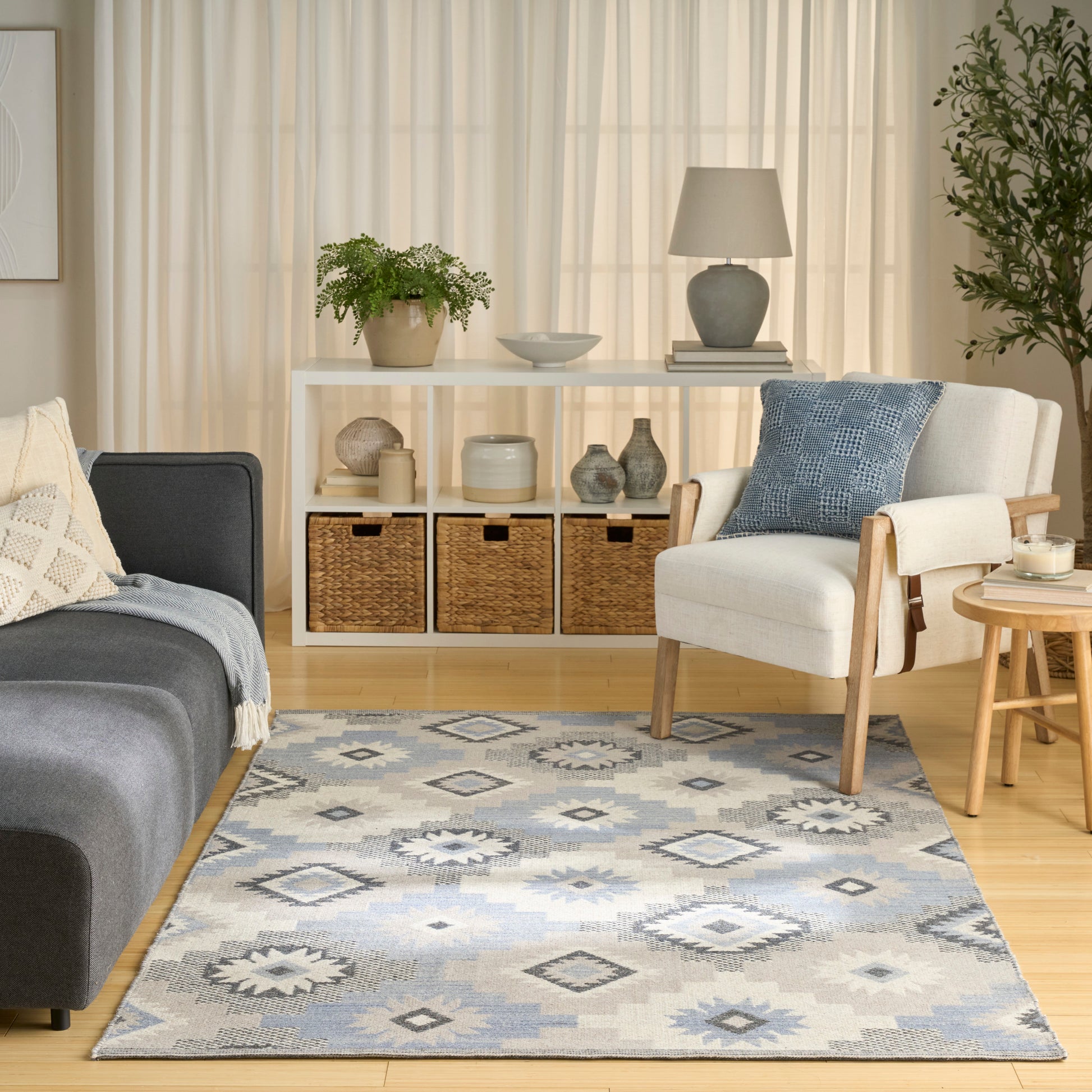 Cozy living room with light wood floors, blue-gray geometric rug, gray sofa, white armchair, woven baskets and greenery.