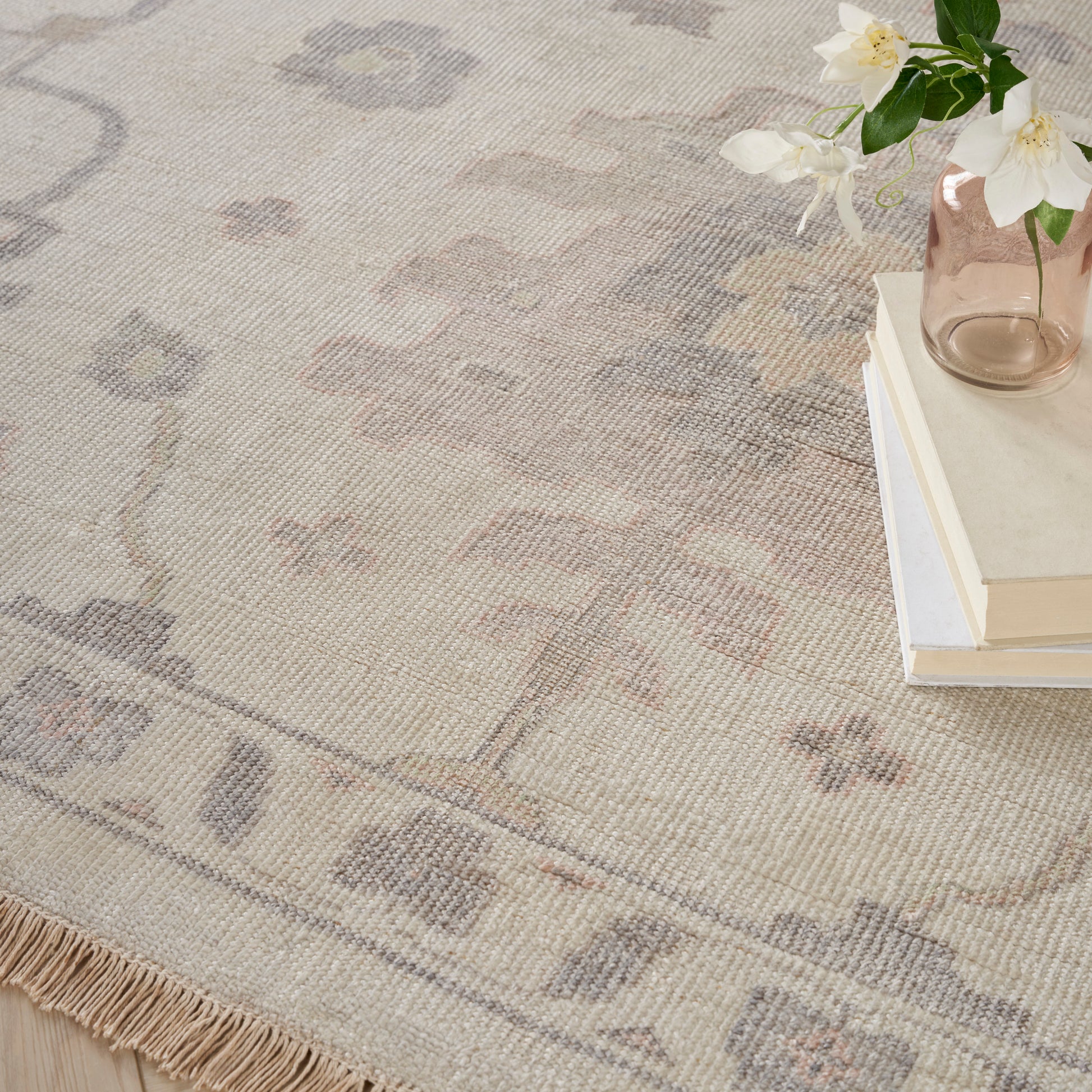 Beige oriental rug with fringe and soft gray-blue motifs, paired with books and a pale pink vase with white flowers.