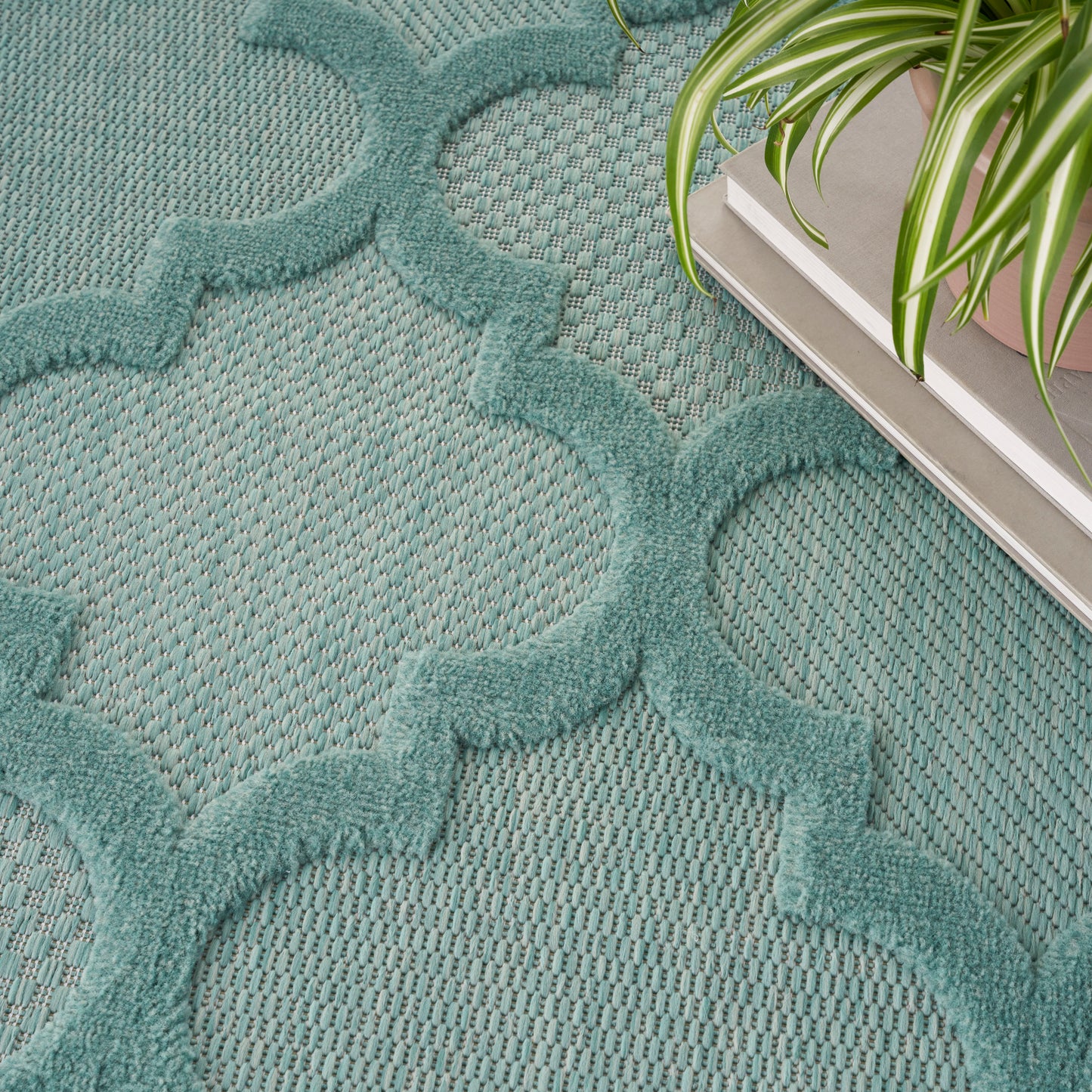 Teal rug with raised quatrefoil lattice pattern on woven base; contemporary decor with a potted plant nearby.