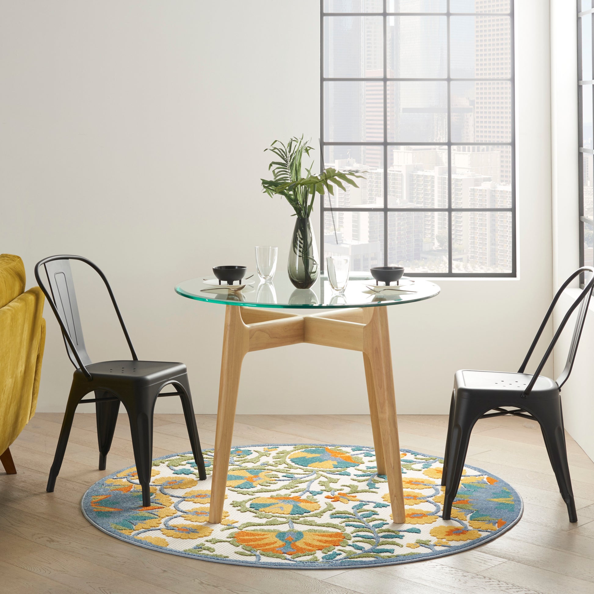 Round glass-top dining table with light wood pedestal, black metal chairs, colorful circular rug, vase of greenery.