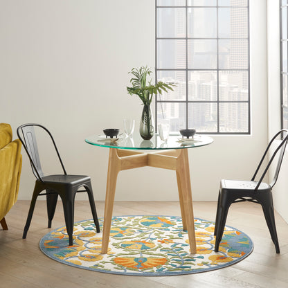 Round glass-top dining table with light wood pedestal, black metal chairs, colorful circular rug, vase of greenery.