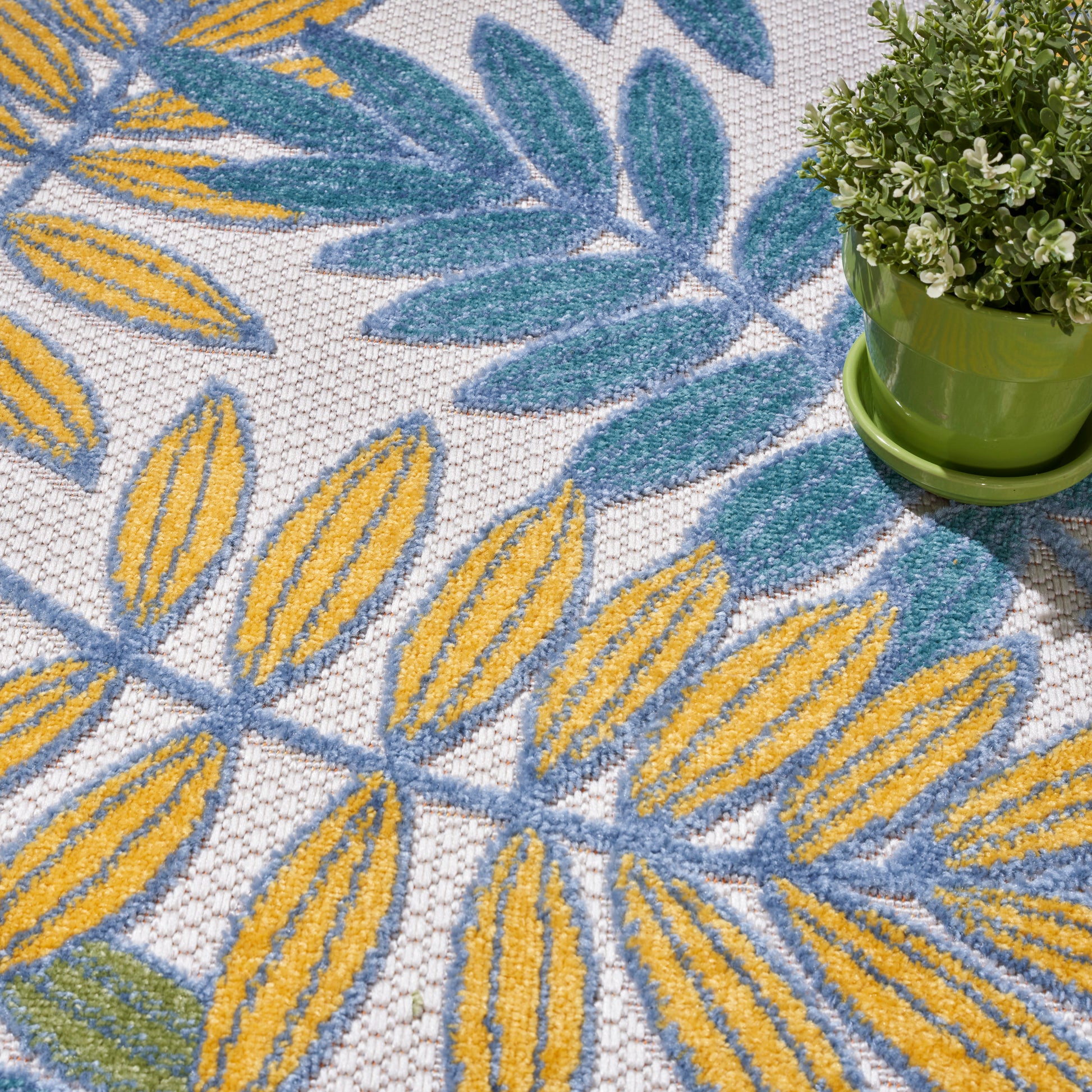 Close-up of a rug with yellow and blue leaf motifs on white, plus a small green plant in a lime-green pot.