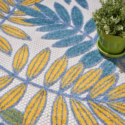 Close-up of a rug with yellow and blue leaf motifs on white, plus a small green plant in a lime-green pot.