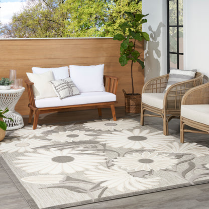 Gray floral outdoor rug (6' x 9') on a patio, surrounded by a wooden sofa with white cushions, wicker chairs, and a potted plant.