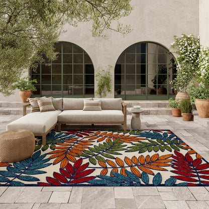 Outdoor patio with beige modular sofa, round wicker ottoman, and a vibrant tropical leafy rug; arched windows and potted plants.
