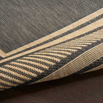 Close-up of a dark charcoal woven rug with beige fringe and striped border on a wooden floor.