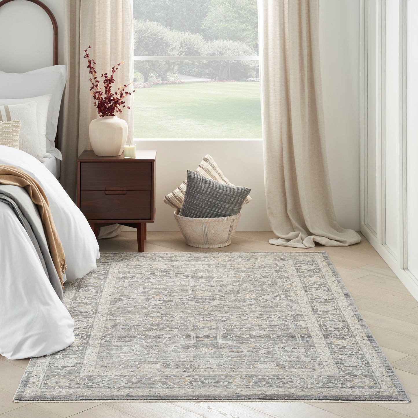 Gray patterned area rug with ornate border in a serene bedroom with a wooden nightstand, beige curtains & wicker basket.