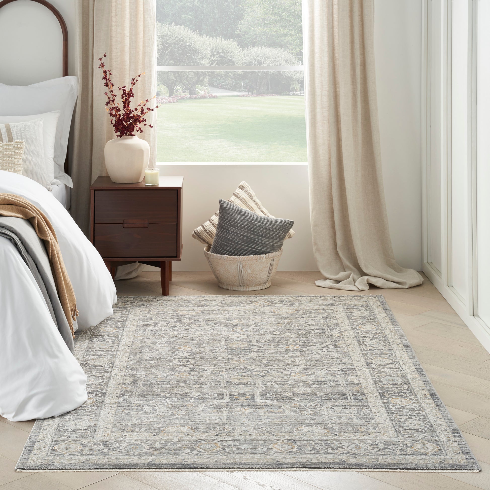 Gray patterned area rug with ornate border in a serene bedroom with a wooden nightstand, beige curtains & wicker basket.