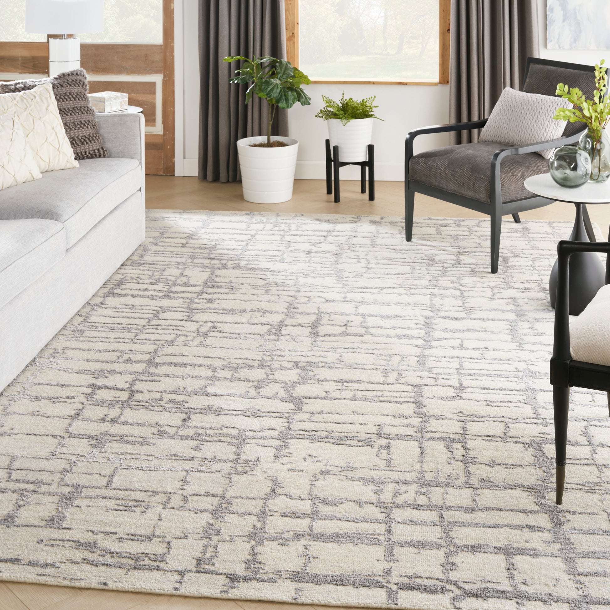 Modern living room: light gray sofa, geometric rug, black metal-framed chair, round side table with vase, plants by window.