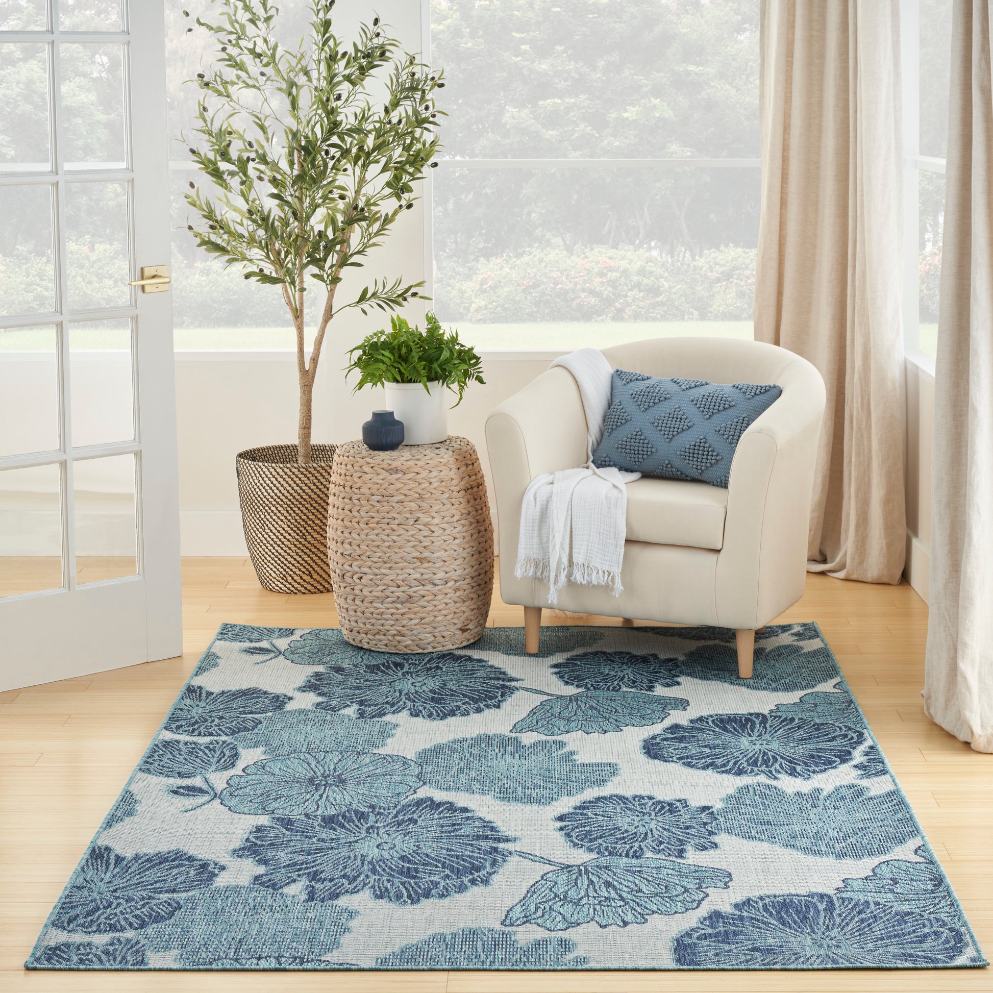 Teal floral area rug under a beige armchair with blue cushion and white throw; woven side table and potted greenery.