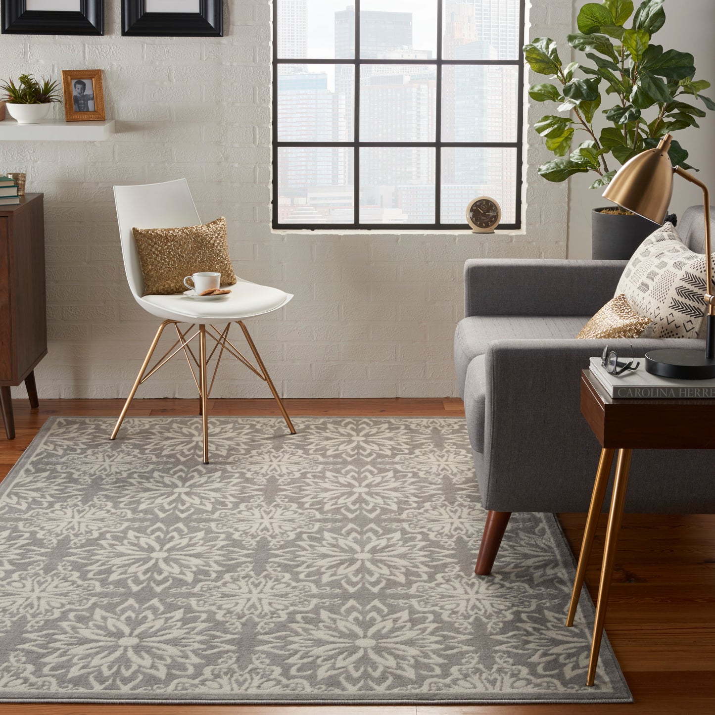 Gray patterned farmhouse rug in a modern living room with a white molded chair, gray sofa, brass lamp, wood side table, plant, and city view.