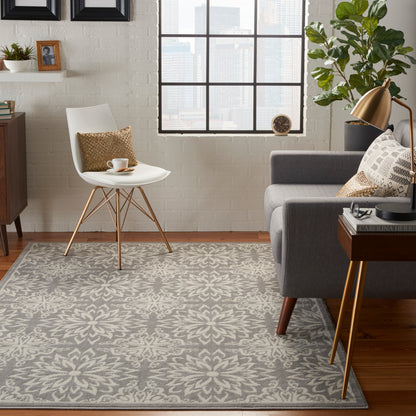 Gray patterned farmhouse rug in a modern living room with a white molded chair, gray sofa, brass lamp, wood side table, plant, and city view.