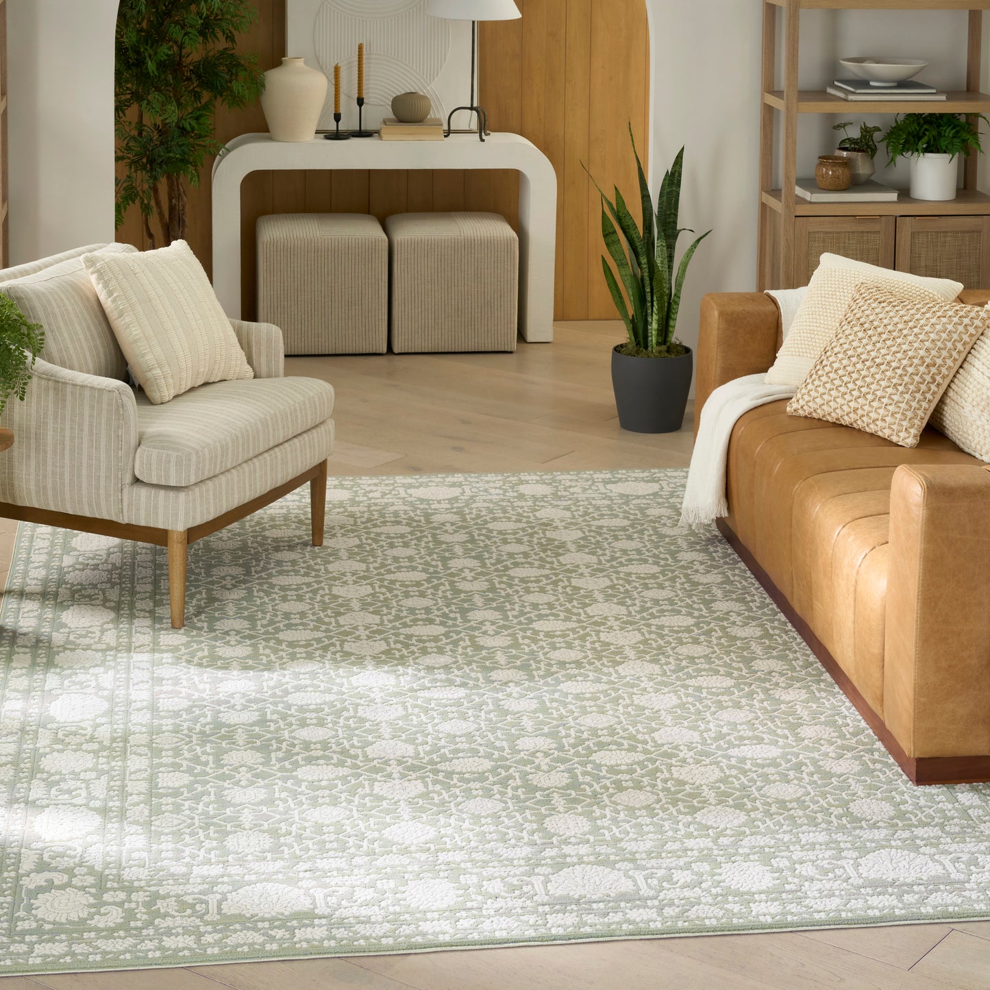Neutral living room with light patterned rug, tan leather sofa, beige armchair on wooden legs, throw pillows & potted plants.