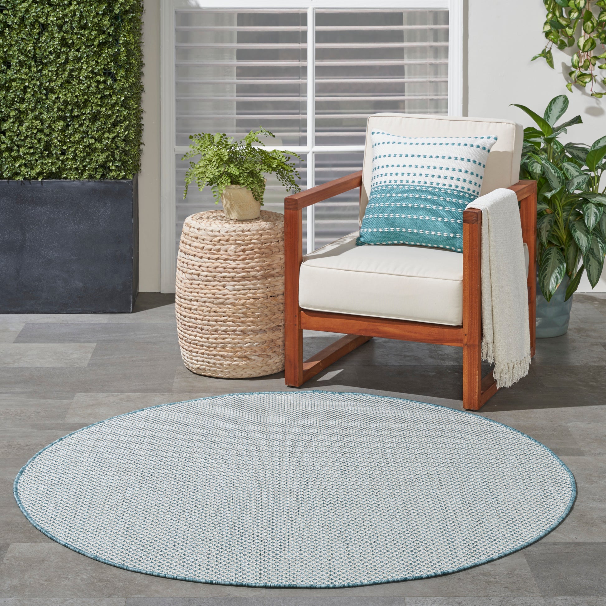 Round light-blue woven outdoor rug; wooden armchair with cream cushion, teal gradient pillow, planter, and draped throw.
