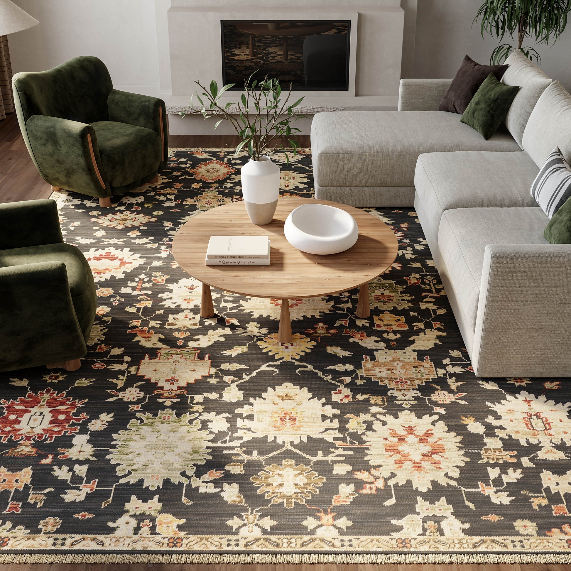 Living room with dark floral rug, round wooden coffee table, gray sectional sofa, green armchairs, fireplace & plant.