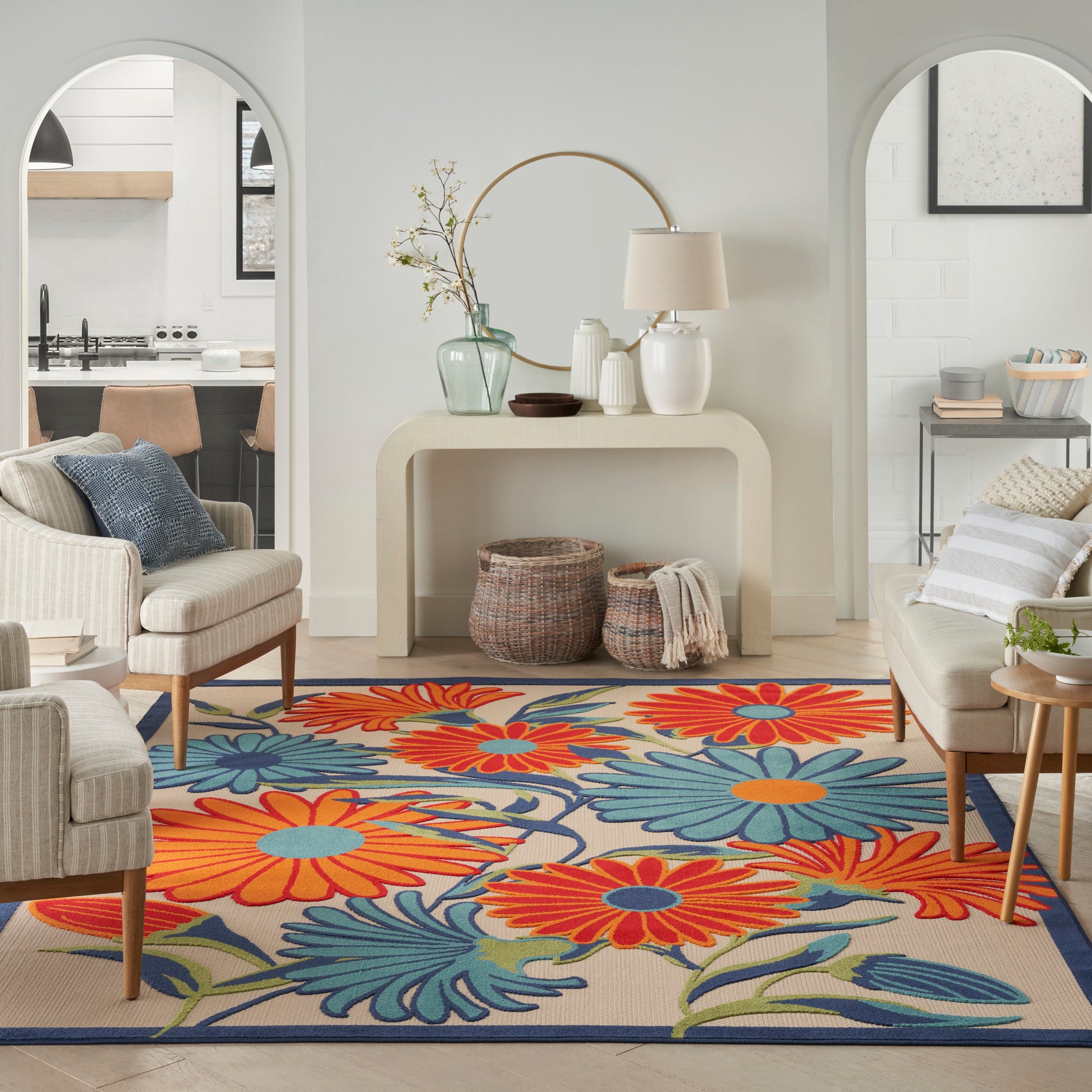 Neutral living room with a bold floral rug in orange, blue and teal, cream seating, and a curved console with a round mirror.