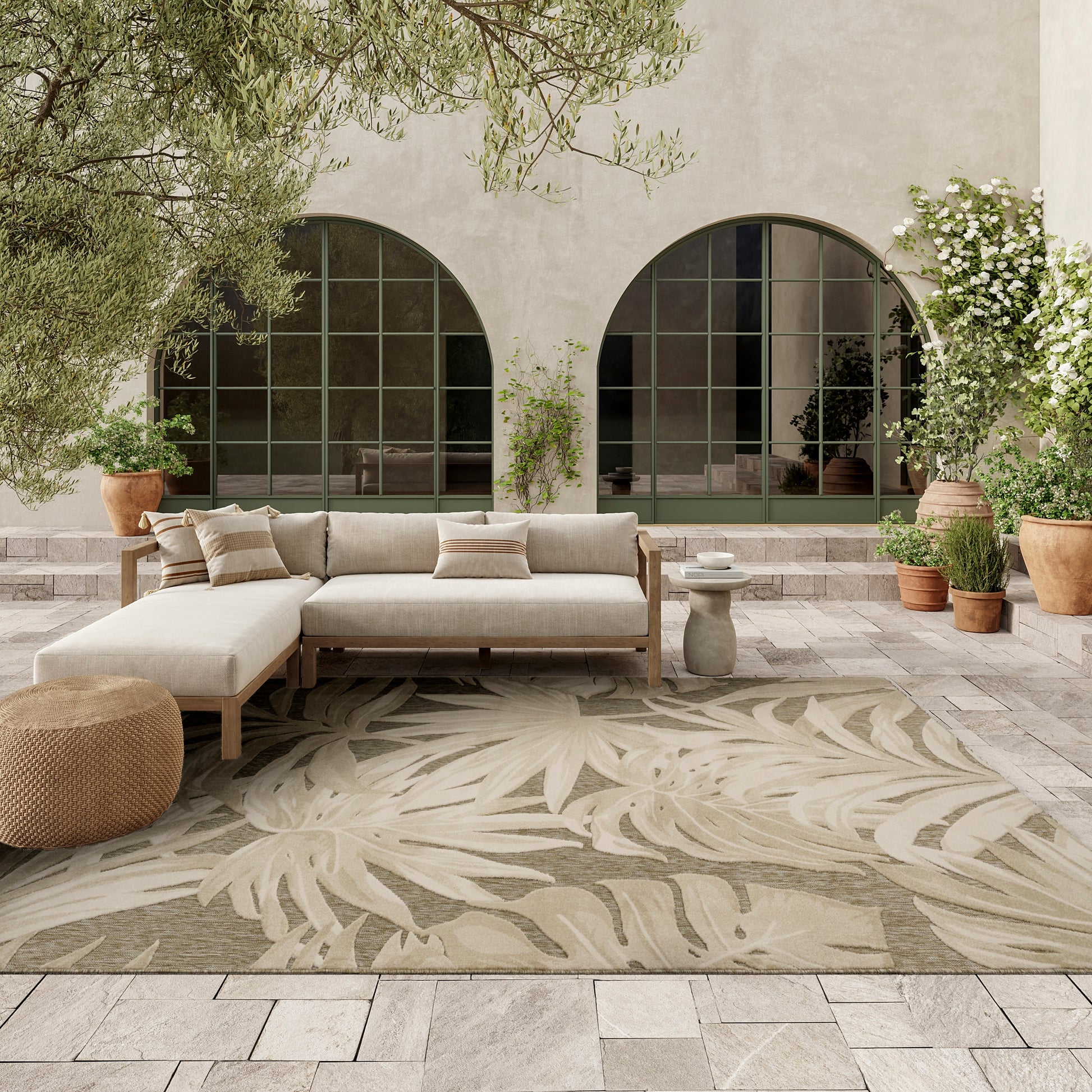 Beige outdoor sectional sofa with a wooden frame on a leaf-pattern rug, arched green-framed windows and potted plants.