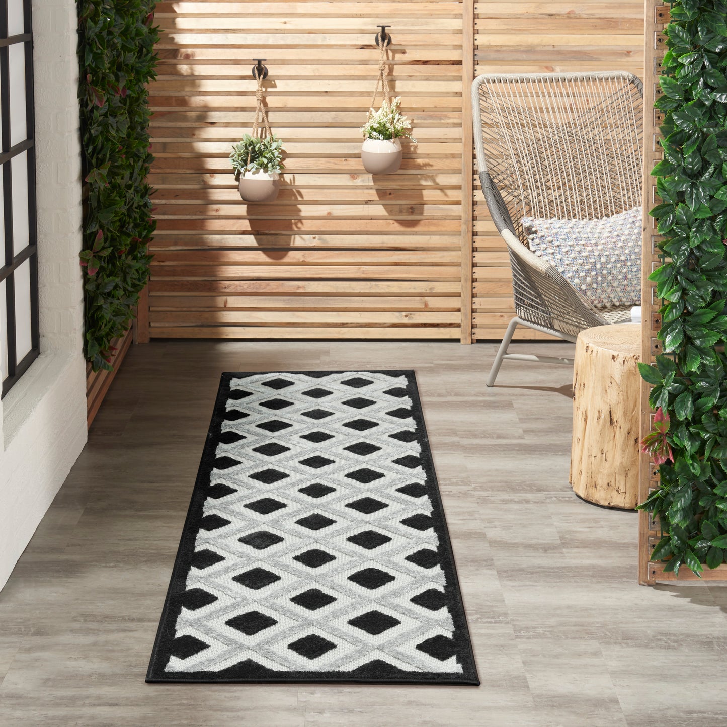 Outdoor balcony runner rug with black border & white diamond pattern, paired with wicker chair & hanging planters.
