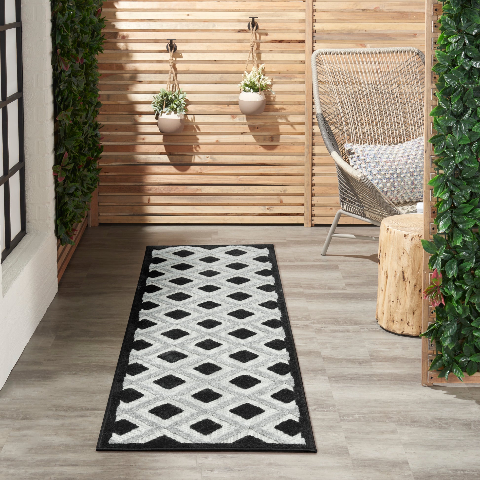 Outdoor balcony runner rug with black border & white diamond pattern, paired with wicker chair & hanging planters.