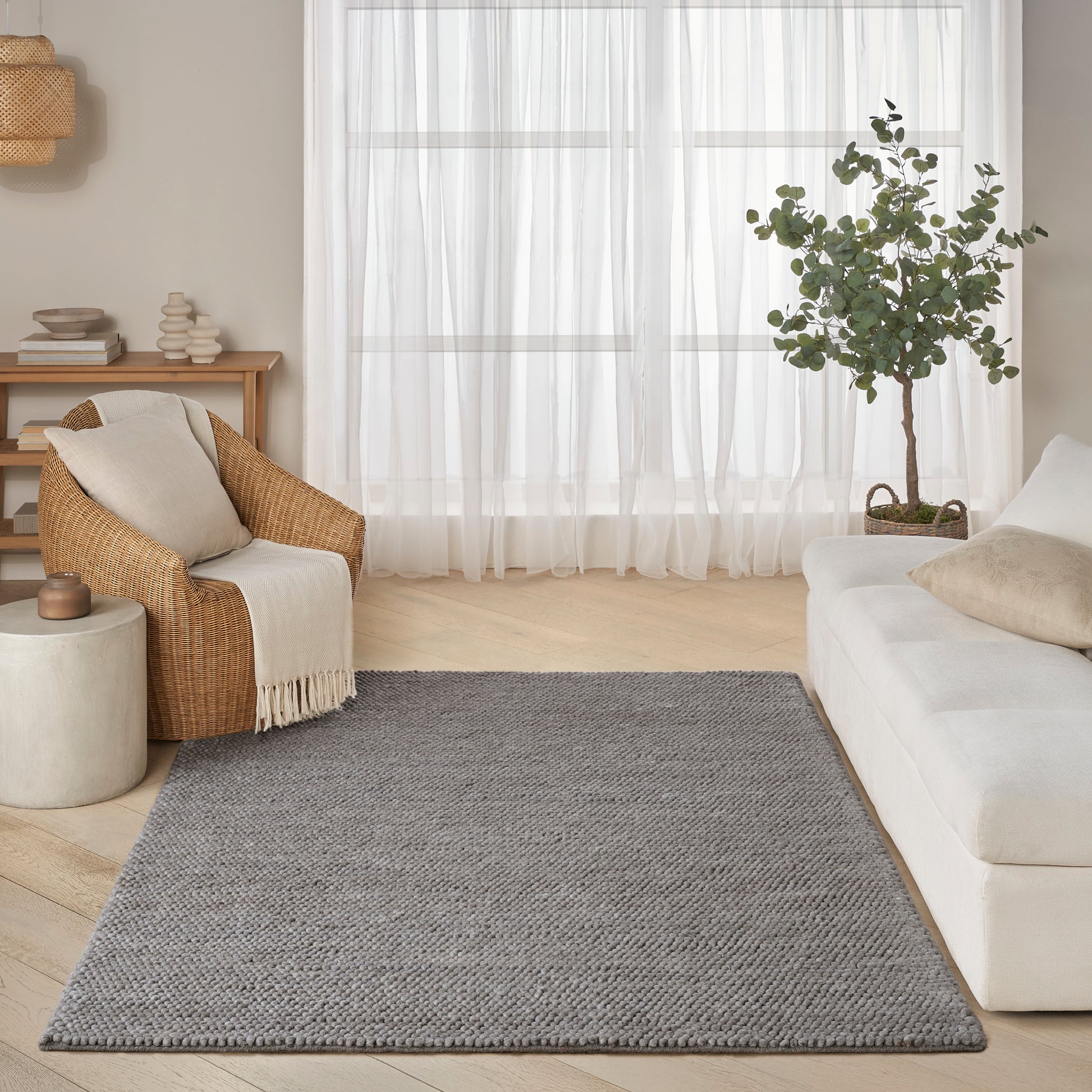 Rectangular grey textured rug in a bright living room with white sofa, wicker chair, sheer curtains, and potted plant.