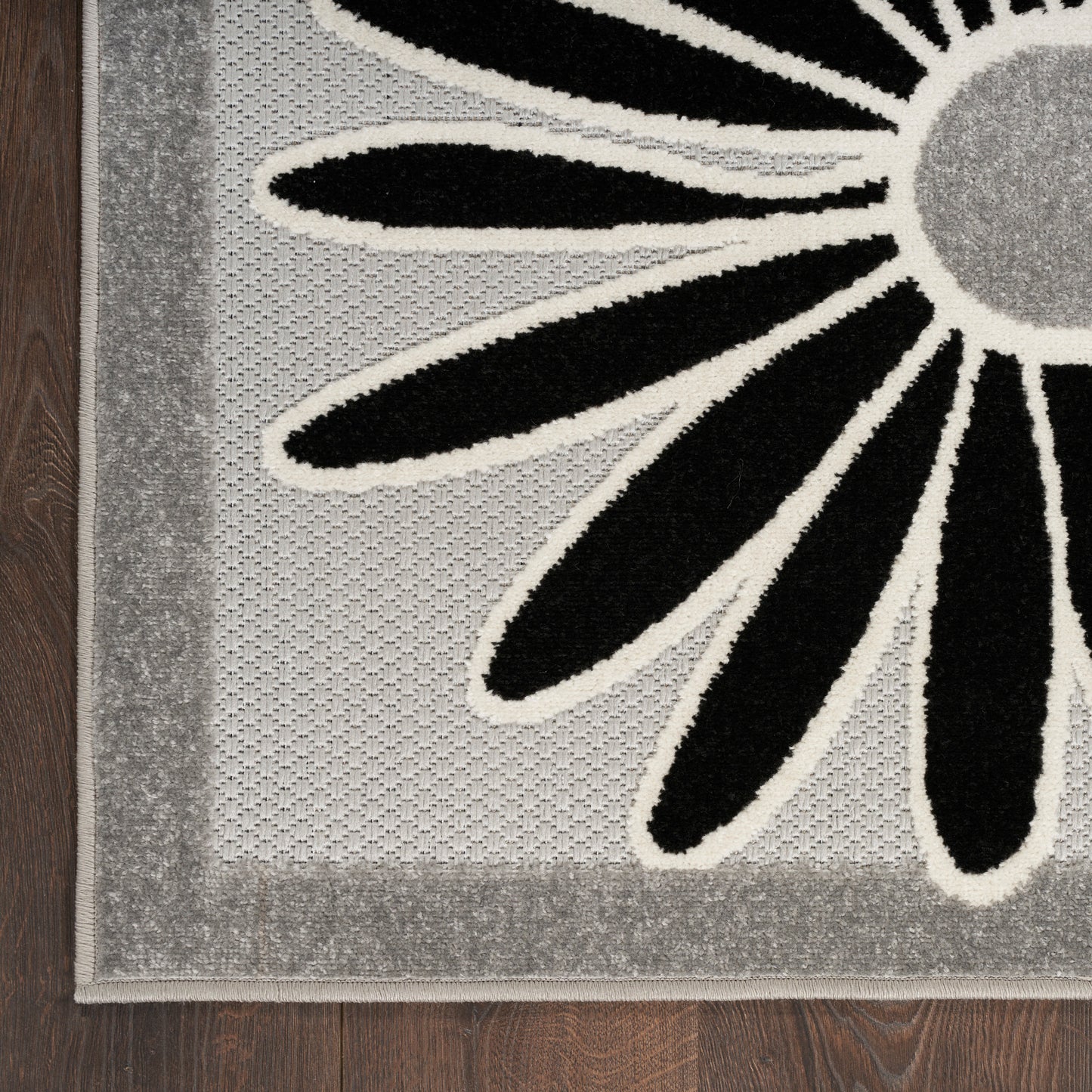 Floral rug with black petals outlined in white on a gray speckled field and pale gray border, on dark wood floor.